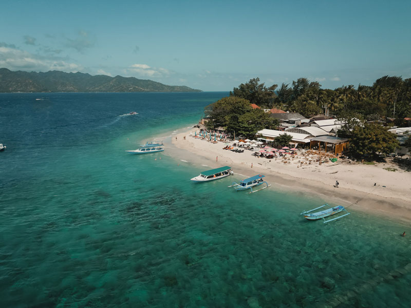 Aerial view of Bahia Gili Air with turquoise water, boats anchored near the beach, and the coastline stretching toward Lombok.