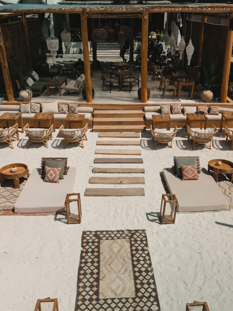 Beachfront seating at Bahia Gili Air with sunbeds, wooden tables, and a tropical open-air restaurant setting on white sand.