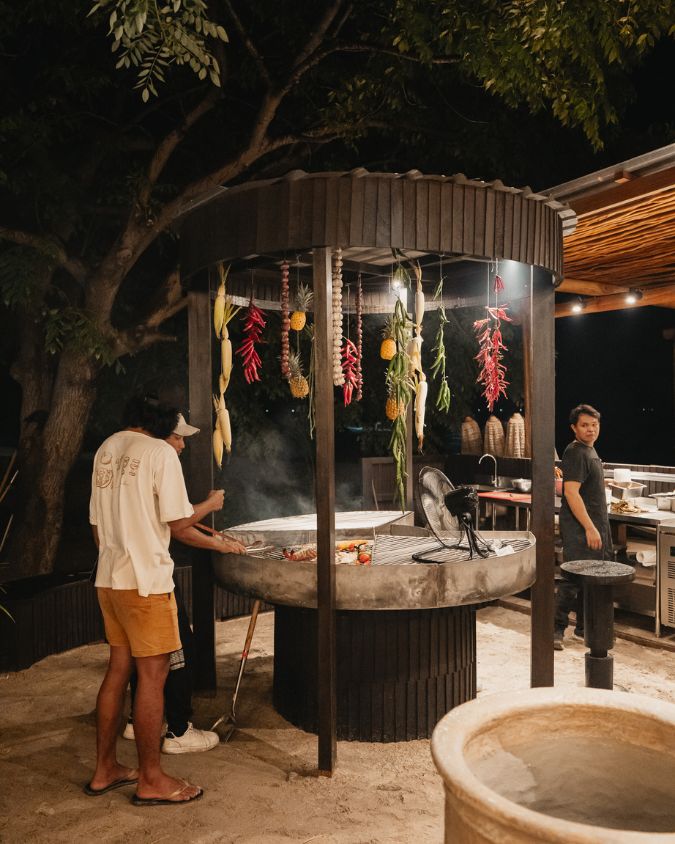A chef cooking fresh skewers and seafood over an outdoor charcoal barbeque station under the stars at BAHIA Gili Air.