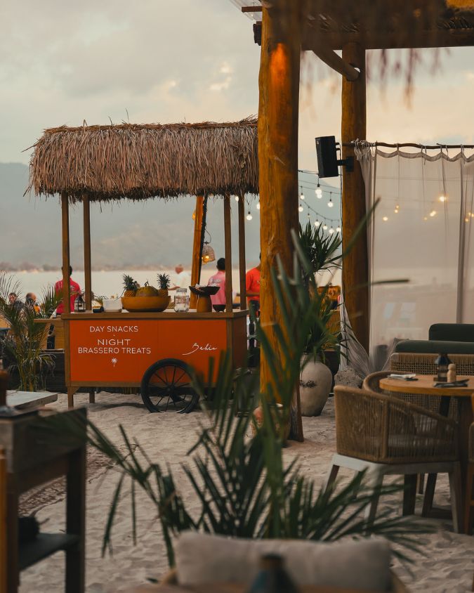 Beachfront tables, brasero snak cart, wicker chairs and tropical plants beside a thatched food cart at Bahia Restaurant & Beachbar in Gili Air.