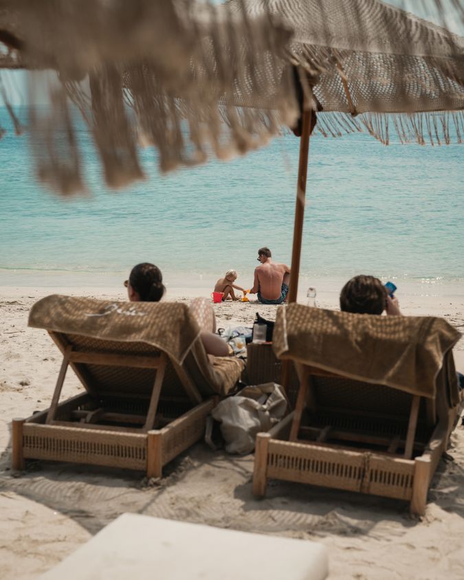 Families relaxing on sunbeds under a thatched canopy and a little kid and his father are playing at the beach Bahia Restaurant & Beachbar, facing the sea in Gili Air.