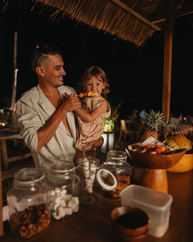 A man and his toddler beside jars of marshmallows at the brasero snack cart at Bahia Restaurant & Beachbar in Gili Air at night.