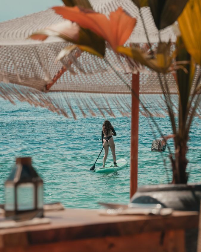 A guest paddleboarding in front of Bahia Restaurant & Beachbar in Gili Air, viewed from the shaded beachfront seating.